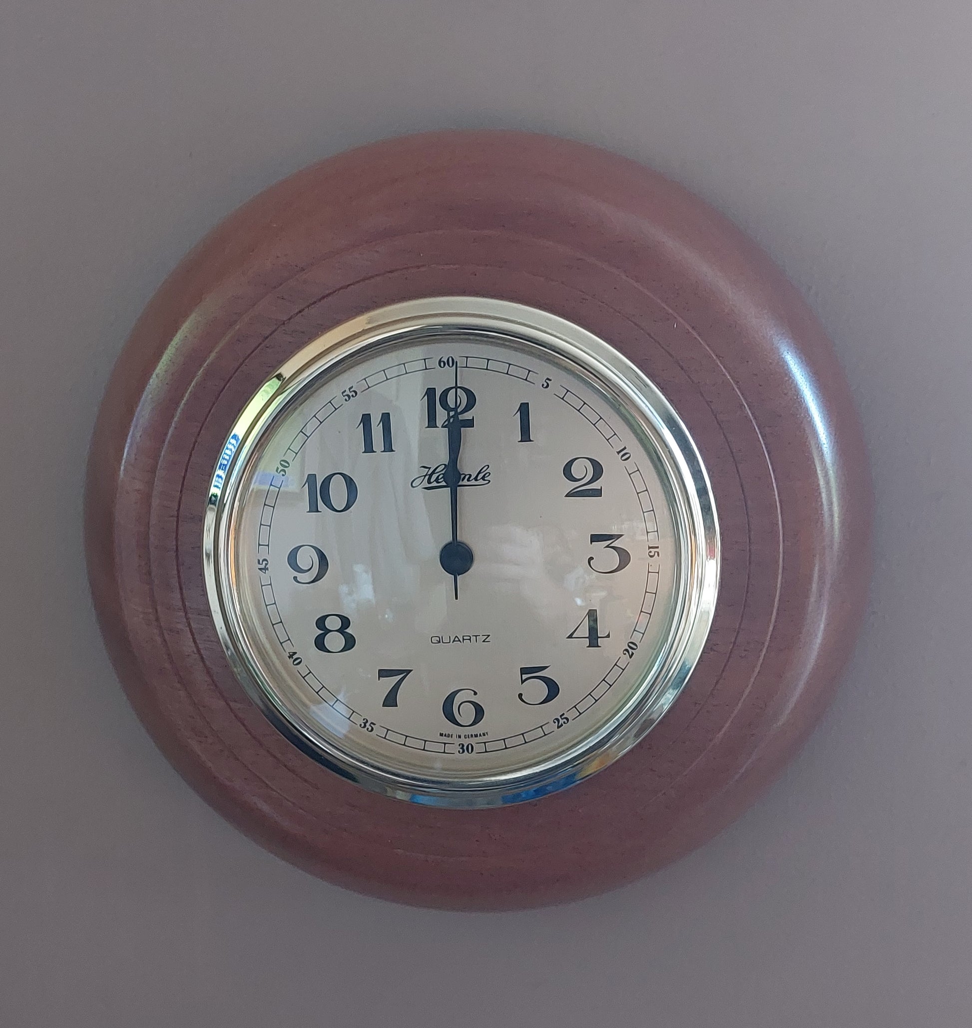 A round wall clock with a reddish-brown wooden frame, a polished silver-tone bezel, and a white dial featuring black Arabic numerals and hands. The brand name 'Amek' is visible on the clock face.