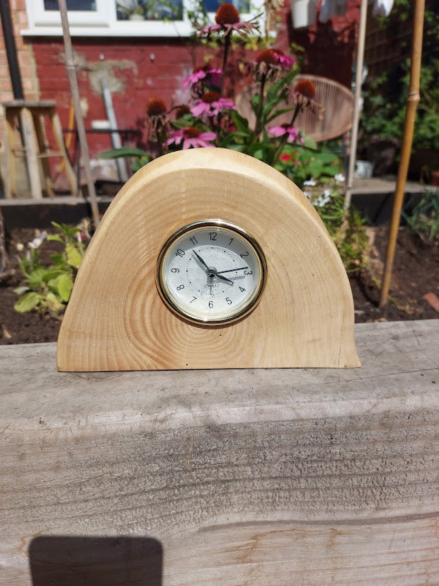 A small wooden mantel clock with a semi-circular natural wood body and a round white clock face, photographed outdoors on a wooden surface with a garden background.