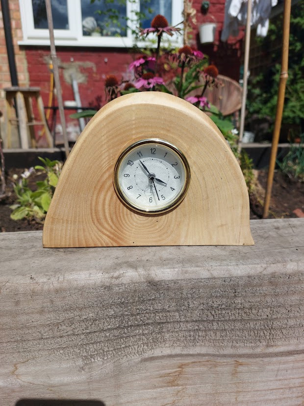 A small wooden mantel clock with a curved natural wood body and a round white clock face, photographed outdoors on a weathered wooden surface with a garden background.