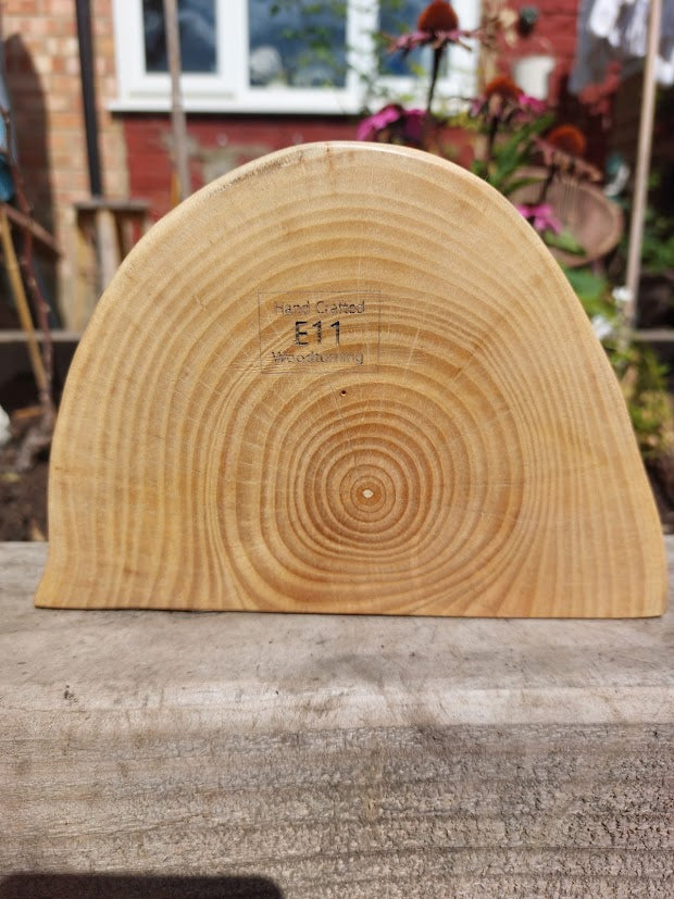 A hand-called natural wood half-moon plaque with visible tree growth rings and the engraving 'Hand Called E11', displayed on a rustic wooden surface with a garden background.