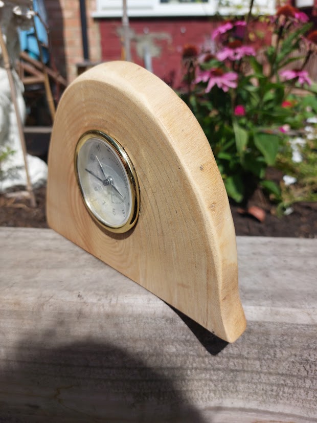 A small wooden mantel clock with a semi-circular light oak body, a brass bezel, and a white dial, photographed outdoors on a weathered wooden surface with garden flowers blurred in the background.